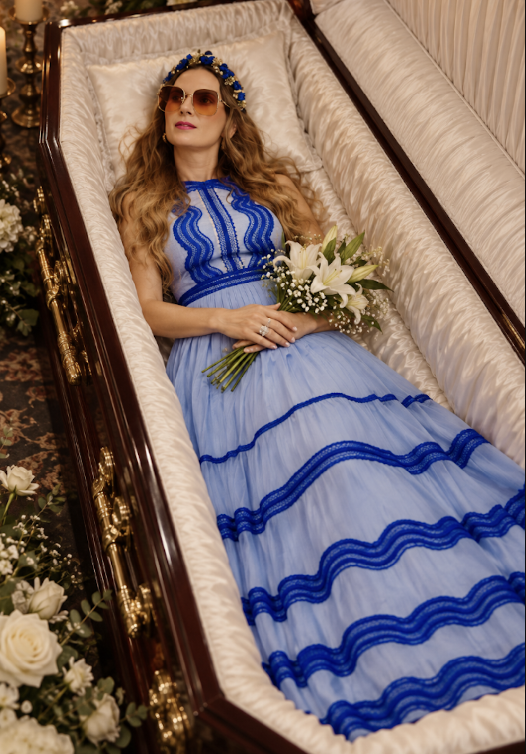 Sahra-Josephine Hjorth lying in an open coffin wearing sunglasses and a blue gown, holding a bouquet of white lilies