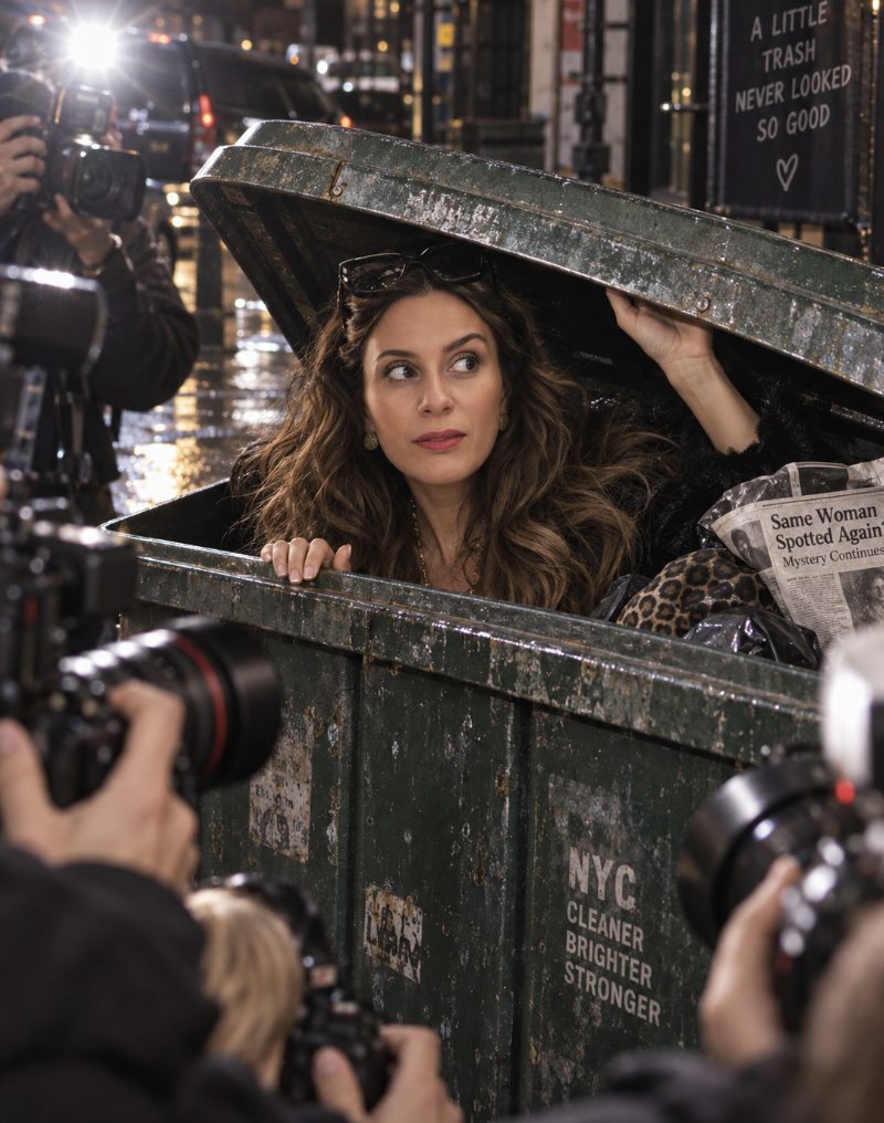 Sahra-Josephine Hjorth emerging from a green NYC dumpster surrounded by photographers, with a newspaper headline reading Same Woman Spotted Again